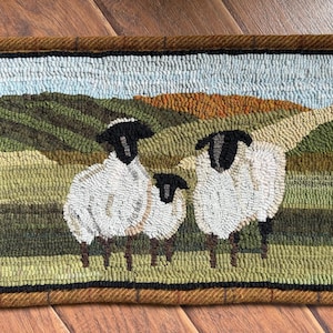 May include: A rectangular hooked rug featuring a landscape scene with three sheep standing in a green field. The sheep have white wool and black faces. The background shows rolling hills in shades of green and brown under a light blue sky.