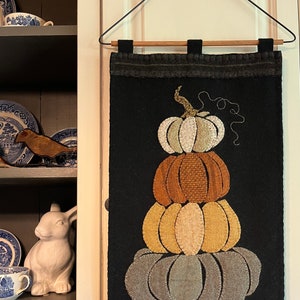 May include: A black felt wall hanging with a stack of five pumpkins in various shades of brown, orange, and beige. The pumpkins are stitched together with a decorative stitch.