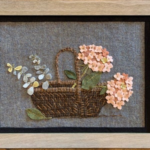 May include: A brown wicker basket filled with pink and white felt flowers. The flowers are arranged in a bouquet and are surrounded by green felt leaves. The basket is sitting on a gray felt background.