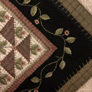 May include: Close-up of a handcrafted textile with a geometric design. The central pattern features brown and white triangles, framed by a reddish-brown border. Black fabric surrounds the design, adorned with embroidered vines and leaves.