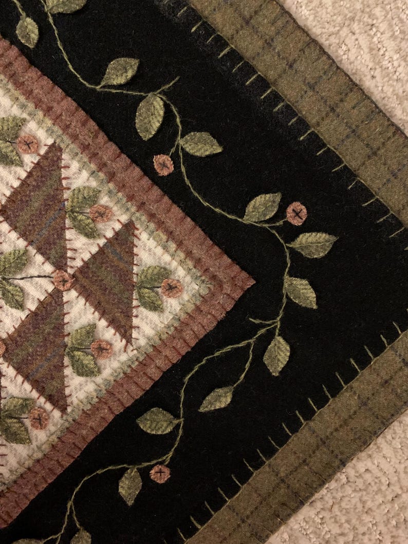 May include: Close-up of a handcrafted textile with a geometric design. The central pattern features brown and white triangles, framed by a reddish-brown border. Black fabric surrounds the design, adorned with embroidered vines and leaves.