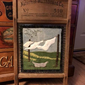 May include: A vintage wooden washboard with a framed needlepoint design depicting a clothesline with laundry hanging on it. The washboard is labeled "National Washboard Co. No. 510 Trade Marks Reg. U.S. Pat. Off. Made in U.S.A. Chicago. Saginaw. Memphis."