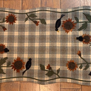 May include: A gray and white plaid rug with a scalloped edge features an embroidered design of black birds and sunflowers.