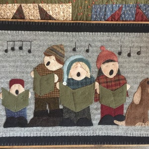 May include: A felt wall hanging with a winter scene of four children singing carols with a dog beside them. The children are wearing winter clothing and holding songbooks. The background is a gray felt with musical notes.