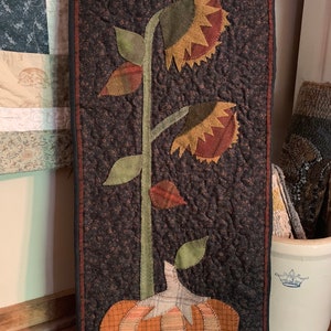 May include: A quilted wall hanging with a brown background features a green stem with two brown and yellow sunflowers and a brown and orange pumpkin at the bottom.