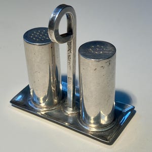 May include: A vintage silver salt and pepper shaker set. The set includes two cylindrical shakers with perforated tops, a rectangular tray, and a handle. The handle is connected to the tray and shakers. The set is marked "LEONARD POSNOW STERLING".