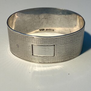 May include: A silver napkin ring with a textured, wave-like pattern. The ring has a rectangular, smooth area with an inscription. The ring is approximately 1.5 inches in diameter and 1 inch tall.