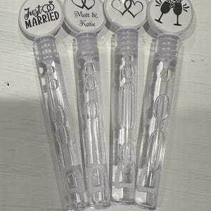 May include: Four clear plastic bubble wands with white labels. The labels have different wedding-themed designs, including a ring, two hearts, two hearts with the text "Matt & Katie", and two champagne glasses with the text "Mr & Mrs".