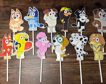 May include: A collection of eleven cartoon dog character cutouts on wooden sticks. The dogs are in various colors, including orange, blue, yellow, pink, and black and white. Some dogs are wearing accessories like glasses and hats. These are likely cake toppers or party decorations.