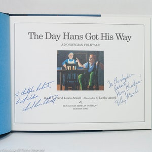 May include: Open book with the title "The Day Hans Got His Way" and a Norwegian folktale. The illustration shows two people at a table. The book has blue covers and handwritten signatures.