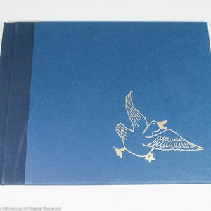 May include: A blue book with a dark blue spine. A gold outline of a duck in flight is on the front cover. The book is lying on a white surface.