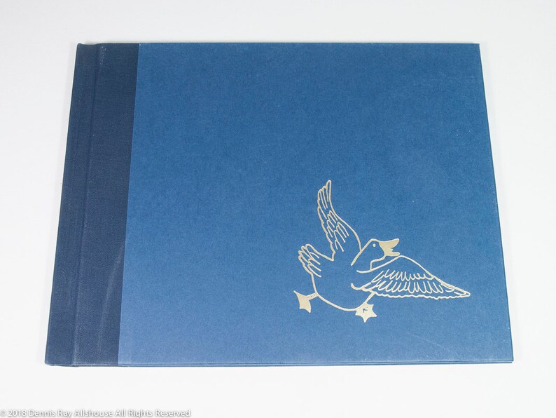 May include: A blue book with a dark blue spine. A gold outline of a duck in flight is on the front cover. The book is lying on a white surface.