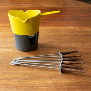 May include: A bright yellow ceramic fondue pot with a heart-shaped rim and a single handle, resting on a dark gray textured base. Beside the pot are six metal fondue forks, each featuring a long, slender shaft and a dark brown wooden handle.