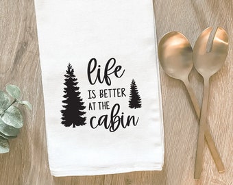 May include: White kitchen towel with black text that reads "life is better at the cabin" and two black silhouette pine trees.