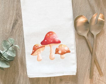 Op de afbeelding: Een witte keukendoek met een rood, oranje en wit paddenstoelenontwerp. De paddenstoelen zijn geschilderd in aquarelstijl.