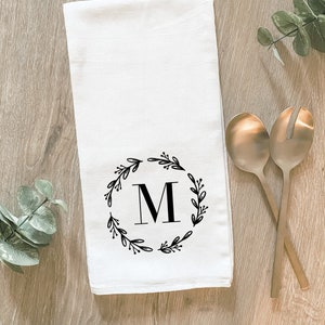 White cotton tea towel with wreath design. In the center of the wreath is an initial. We can print any initial you would like. Design is printed with black ink.