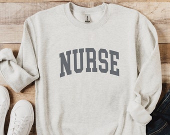 Puede incluir: Una sudadera gris claro con la palabra "NURSE" impresa en gris oscuro en la parte delantera.