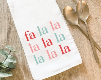 May include: White kitchen towel with the words "fa la la" in red, pink, and teal lettering. The towel is folded and lies on a wooden surface, with gold serving utensils in the background.