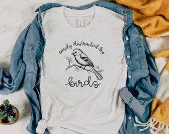 Może przedstawiać: Biała koszulka z czarno-białym rysunkiem ptaka na gałęzi. Na koszulce nadrukowany jest tekst "easily distracted by birds".