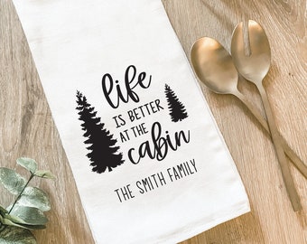 May include: White kitchen towel with black text that reads "life is better at the cabin The Smith Family" with two black pine tree silhouettes.