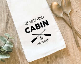 May include: White kitchen towel with black text reading "THE SMITH FAMILY CABIN" featuring crossed oars and an anchor graphic. Below the graphic is the text "LAKE MONONA". Two gold serving spoons are on the right.