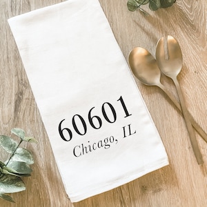 May include: White kitchen towel with the text "60601 Chicago, IL" printed in black. Two gold-colored serving spoons are on the right side of the towel. The towel is on a light brown wooden surface.