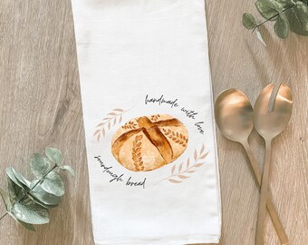 May include: White kitchen towel with a watercolor illustration of a sourdough bread loaf. The towel has the words "handmade with love" and "sourdough bread" printed on it. Two gold serving spoons are next to the towel.