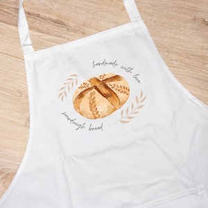 May include: White apron featuring a watercolor illustration of a sourdough bread loaf. The design includes the text "handmade with love" and "sourdough bread" in a handwritten style, surrounded by wheat stalks.