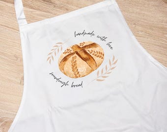 Puede incluir: Delantal blanco con una ilustración de acuarela de una hogaza de pan de masa madre. El diseño incluye el texto "handmade with love" y "sourdough bread" en un estilo manuscrito, rodeado de espigas de trigo.