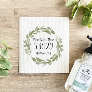 Puede incluir: Toalla de cocina blanca con un diseño de corona de acuarela verde. El texto "Home Sweet Home 53029 Hartland, WI" está impreso en negro dentro de la corona.