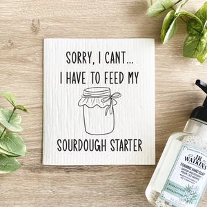 May include: White square sign with the text "SORRY, I CAN'T... I HAVE TO FEED MY SOURDOUGH STARTER" in black. A line drawing of a jar with a tied cloth lid is below the text. A bottle of foaming hand soap is in the background.