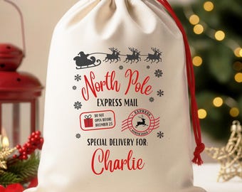 May include: A cream-colored drawstring gift bag with a red drawstring. The bag features a black graphic of Santa's sleigh and reindeer, along with the words "North Pole Express Mail" and "Special Delivery For: Charlie" in red.