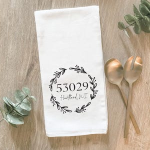 Puede incluir: Toalla de cocina blanca con un diseño de corona floral negra y el texto "53029 Hartland, WI".