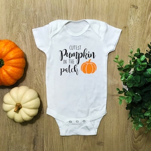 Könnte beinhalten: Weißer Baby-Strampler mit schwarzem Text "Cutest Pumpkin in the Patch" und einer orangefarbenen Kürbisgrafik.