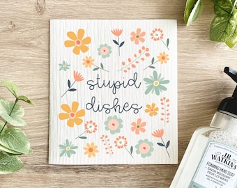 Könnte beinhalten: Ein weißes Geschirrtuch mit dem Schriftzug "stupid dishes" in einer verspielten Schriftart, umgeben von bunten Blumenillustrationen in Orange, Gelb und Türkis. Im Hintergrund ist eine Flasche Schaumhandseife zu sehen.