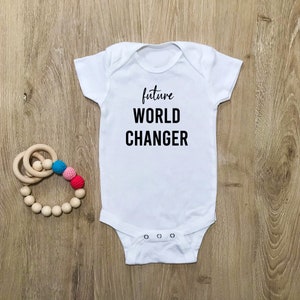 Puede incluir: Body de bebé blanco con el texto negro "future WORLD CHANGER". Un sonajero de madera con cuentas de colores está al lado del body. El body tiene un cierre de broches.