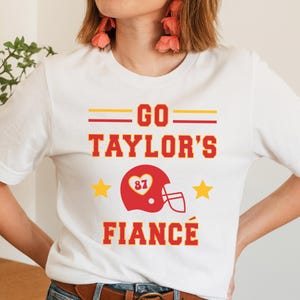 Puede incluir: Camiseta blanca con texto rojo y amarillo que dice "GO TAYLOR'S FIANCÉ". El diseño incluye un casco de fútbol americano rojo con el número 87 dentro de un corazón y dos estrellas amarillas.