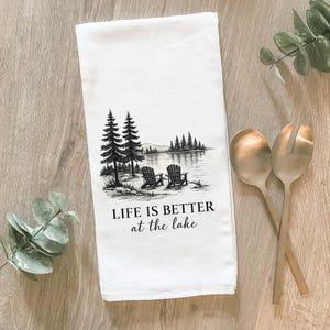 May include: A white kitchen towel featuring a black ink illustration of two Adirondack chairs on a lakeshore with pine trees and the text "LIFE IS BETTER at the lake" printed in a combination of bold sans-serif and elegant script fonts.