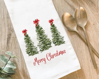 Peut inclure: Torchon de cuisine blanc sur le thème de Noël, avec trois sapins verts ornés de nœuds rouges. Les mots "Merry Christmas" sont imprimés en rouge en dessous. Le torchon est en matière douce et absorbante.