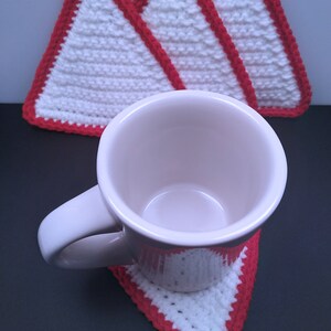 May include: Three white crocheted triangular coasters with red trim, one of which is holding a white ceramic mug.