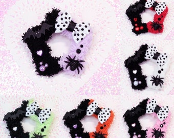 May include: Six star-shaped hair scrunchies with different color combinations. Each scrunchie has a black and white polka dot bow, a black bat, a black spider, and black and white accents. The scrunchies are made of fuzzy fabric.