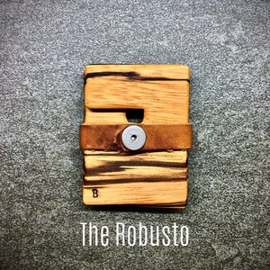 May include: A wooden wallet with a brown leather strap and a metal button. The wallet has a wood grain pattern and the text "The Robusto" is printed below it.
