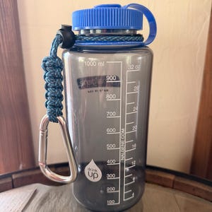 May include: A translucent gray Nalgene water bottle with a blue screw-top lid and a blue paracord handle attached to a silver carabiner. The bottle features white measurement markings in milliliters and ounces, along with a drink up logo.