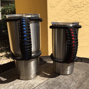 May include: Two stainless steel tumblers with black paracord handles. The handle on the left tumbler is blue and the handle on the right tumbler is red. Both tumblers have the word "YETI" printed on the bottom.