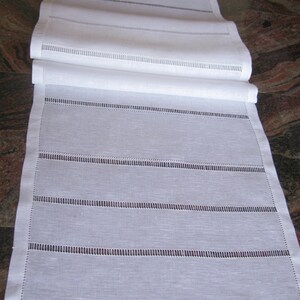 May include: White linen table runner with delicate embroidered lines. The runner is folded in half and has a subtle sheen.