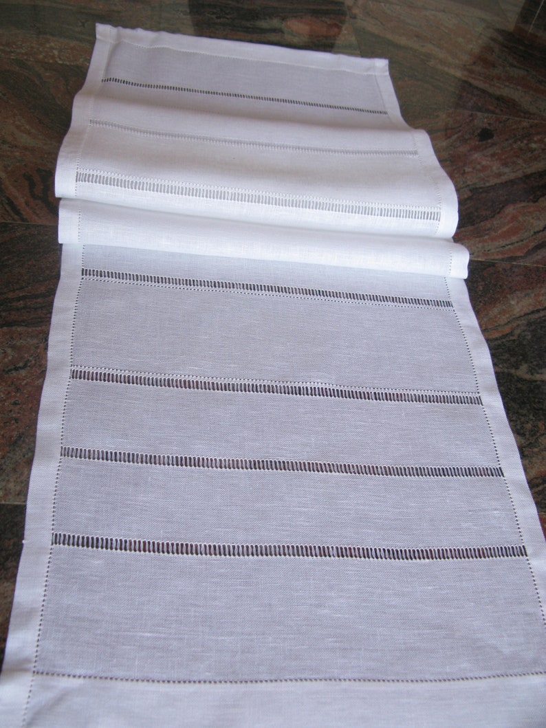 May include: White linen table runner with delicate embroidered lines. The runner is folded in half and has a subtle sheen.