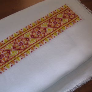 May include: White linen tablecloth with a red, orange and yellow embroidered border. The border features a geometric pattern with repeating squares and floral motifs.