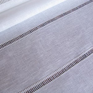 May include: White linen fabric with a subtle texture and a decorative stitched border. The stitching creates a series of parallel lines across the fabric.