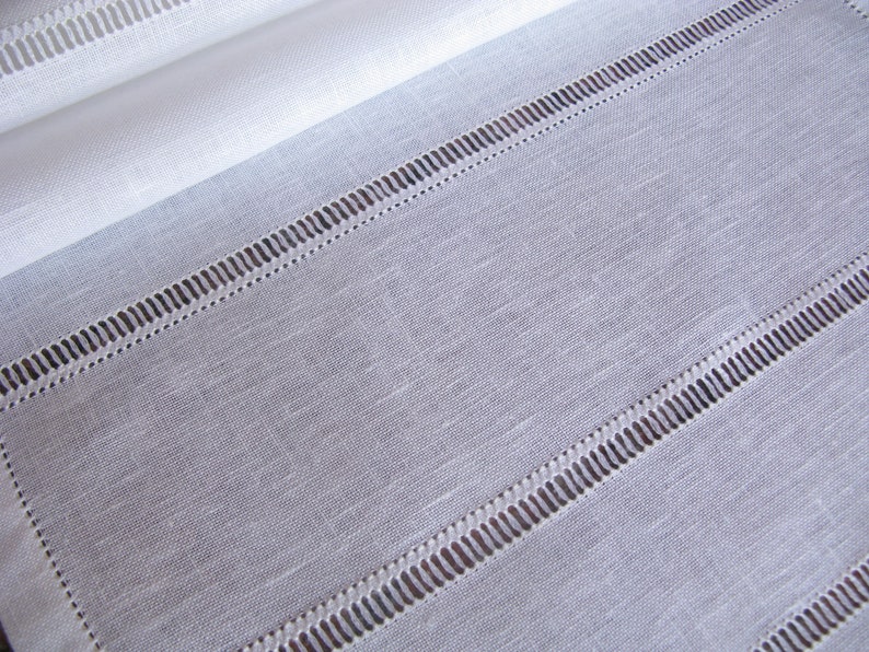 May include: White linen fabric with a subtle texture and a decorative stitched border. The stitching creates a series of parallel lines across the fabric.