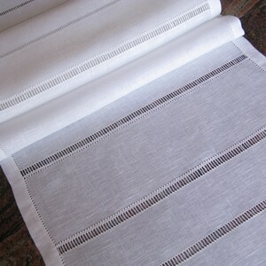May include: White linen table runner with a delicate stitched design. The runner has a subtle texture and a simple, elegant look.